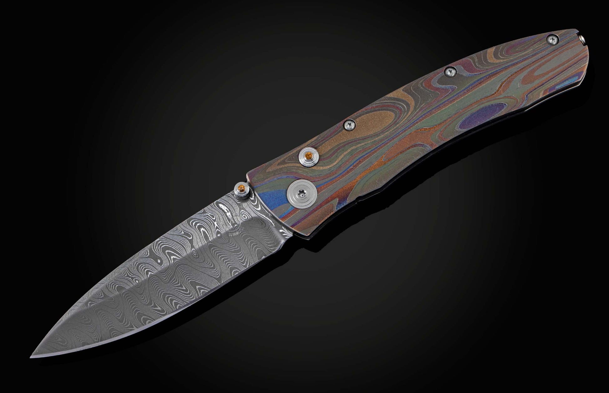 Handcrafted knife from William Henry featuring a hand forged Damascus blade and a titanium frame that is laser-engraved in a swirl of color. The button lock and thumb stud are inset with citrine gemstones. The pocket knife is shown open.