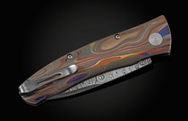 Handcrafted knife from William Henry featuring a hand forged Damascus blade and a titanium frame that is laser-engraved in a swirl of color. The button lock and thumb stud are inset with citrine gemstones. The pocket knife is shown closed.