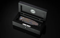 Handcrafted knife from William Henry featuring a hand forged Damascus blade and a titanium frame that is laser-engraved in a swirl of color. The button lock and thumb stud are inset with citrine gemstones. The pocket knife is shown in a custom display cas